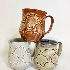 Puede incluir: Tres tazas de cerámica con un diseño festoneado. Las tazas están apiladas una encima de la otra. La taza de arriba es marrón, la taza del medio es blanca y la taza de abajo es verde.