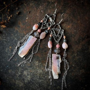 Op de afbeelding: Een paar handgemaakte oorbellen met een zilveren geometrisch ontwerp, roze en grijze stenen en bungelende zilveren kettingen. De oorbellen hebben een unieke, bohemien stijl met een natuurlijke, aardse uitstraling. De stenen variëren in vorm en grootte.