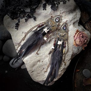 Puede incluir: Un par de pendientes hechos a mano con plumas oscuras, huesos pequeños y cuentas rojas. Los pendientes tienen un diseño de metal color latón vintage con una piedra única en forma de ojo. Los pendientes se exhiben sobre una superficie de piedra clara.