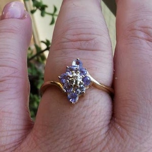 Puede incluir: Un anillo de oro con un grupo de piedras preciosas púrpuras y una sola piedra blanca en el centro.