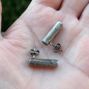 May include: A pair of silver-colored stud earrings. Each earring features a textured, cylindrical design. The earrings are displayed against a blurred green background.