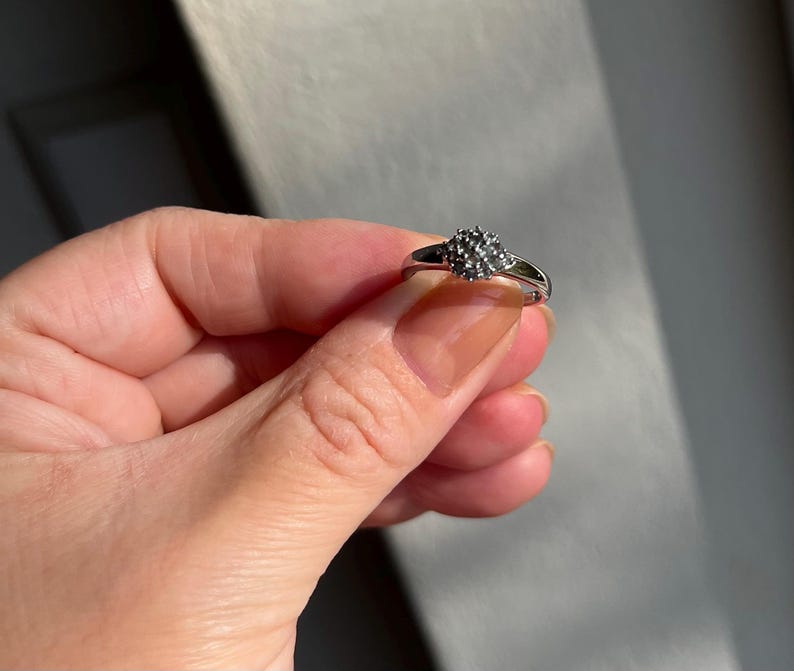 Puede incluir: Un anillo plateado con un grupo de peque&ntilde;as piedras oscuras en el centro. El anillo tiene una banda delgada y pulida, y las piedras est&aacute;n dispuestas en forma redondeada. El anillo se muestra sobre un fondo neutro.