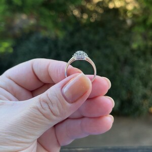 Puede incluir: Un anillo de plata con un grupo de peque&ntilde;as piedras preciosas transparentes en el centro. El anillo se sostiene contra un fondo verde borroso, mostrando su delicado dise&ntilde;o y brillo. El anillo tiene una banda delgada y elegante.