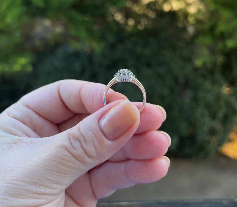 Puede incluir: Un anillo de plata con un grupo de peque&ntilde;as piedras preciosas transparentes en el centro. El anillo se sostiene contra un fondo verde borroso, mostrando su delicado dise&ntilde;o y brillo. El anillo tiene una banda delgada y elegante.
