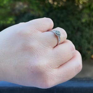 Puede incluir: Un anillo de plata con un grupo de peque&ntilde;as piedras preciosas redondas. El anillo est&aacute; engastado en una banda delgada y se sostiene en una mano contra un fondo verde borroso.