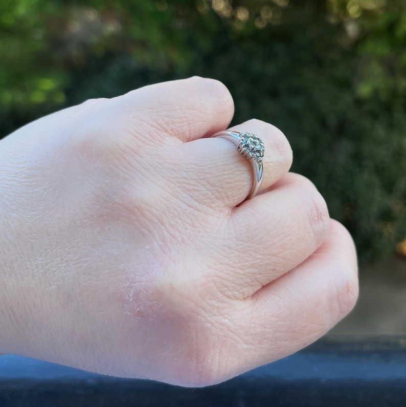 Puede incluir: Un anillo de plata con un grupo de peque&ntilde;as piedras preciosas redondas. El anillo est&aacute; engastado en una banda delgada y se sostiene en una mano contra un fondo verde borroso.