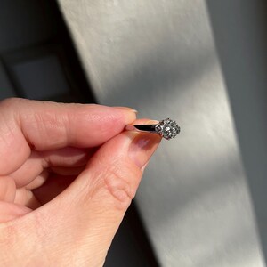 Puede incluir: Un anillo plateado con un grupo de peque&ntilde;as piedras redondas y transparentes que forman un dise&ntilde;o floral. El anillo se sostiene en una mano, centr&aacute;ndose en los detalles del anillo y la luz que reflejan las piedras.