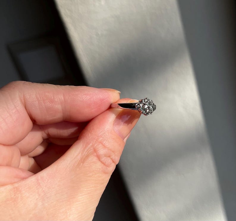 Puede incluir: Un anillo plateado con un grupo de peque&ntilde;as piedras redondas y transparentes que forman un dise&ntilde;o floral. El anillo se sostiene en una mano, centr&aacute;ndose en los detalles del anillo y la luz que reflejan las piedras.
