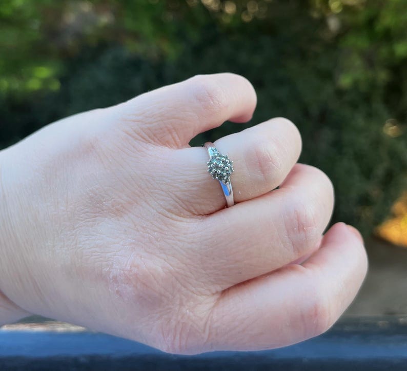 Puede incluir: Un anillo plateado con un grupo de peque&ntilde;as piedras grises redondas. El anillo est&aacute; en un dedo, con la mano enfocada contra un fondo verde borroso. La banda del anillo es delgada y pulida.