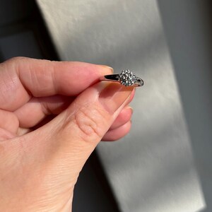 Puede incluir: Un anillo plateado con un grupo de peque&ntilde;as piedras redondas y transparentes. El anillo se sostiene en una mano, con la banda y las piedras captando la luz. El dise&ntilde;o del anillo es de estilo cl&aacute;sico y elegante.