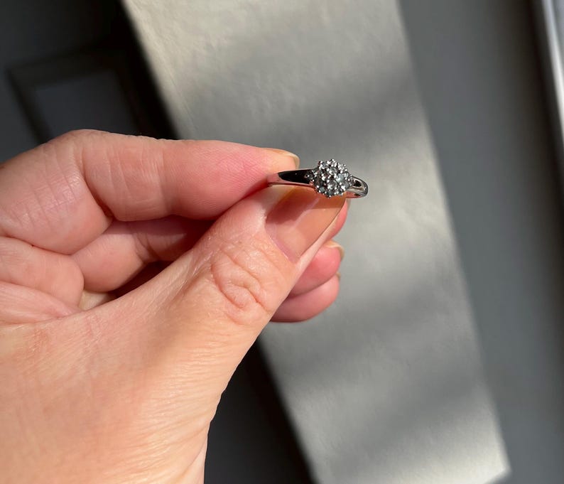 Puede incluir: Un anillo plateado con un grupo de peque&ntilde;as piedras redondas y transparentes. El anillo se sostiene en una mano, con la banda y las piedras captando la luz. El dise&ntilde;o del anillo es de estilo cl&aacute;sico y elegante.