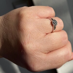 Puede incluir: Un anillo plateado con un grupo de peque&ntilde;as piedras claras dispuestas en un dise&ntilde;o floral o de estrella. El anillo est&aacute; en un dedo, mostrando los intrincados detalles del engaste y el brillo de las piedras. La banda del anillo es delgada y pulida.