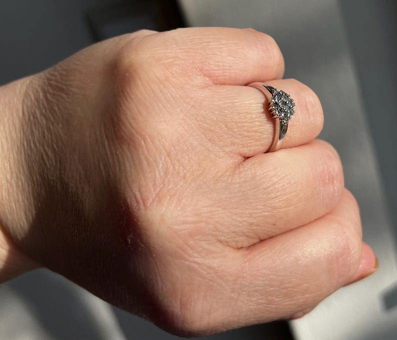 Puede incluir: Un anillo plateado con un grupo de peque&ntilde;as piedras claras dispuestas en un dise&ntilde;o floral o de estrella. El anillo est&aacute; en un dedo, mostrando los intrincados detalles del engaste y el brillo de las piedras. La banda del anillo es delgada y pulida.