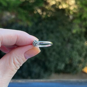 Puede incluir: Un anillo de plata con un grupo de peque&ntilde;as piedras texturizadas. El anillo se sostiene en una mano, con un fondo verde borroso. El dise&ntilde;o del anillo es delicado, destacando su artesan&iacute;a y detalle.