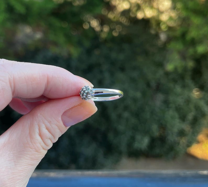 Puede incluir: Un anillo de plata con un grupo de peque&ntilde;as piedras texturizadas. El anillo se sostiene en una mano, con un fondo verde borroso. El dise&ntilde;o del anillo es delicado, destacando su artesan&iacute;a y detalle.