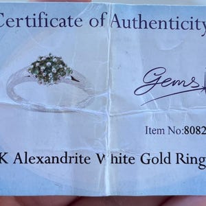 Puede incluir: Un certificado de autenticidad para un anillo de oro blanco de 9K Alexandrita. La tarjeta presenta una imagen del anillo con un grupo de piedras preciosas. El texto incluye "Gems TV" y el n&uacute;mero de art&iacute;culo 80821.