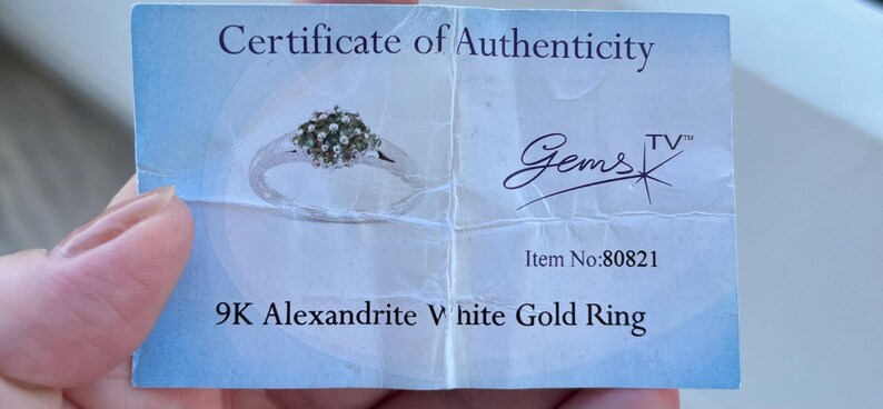 Puede incluir: Un certificado de autenticidad para un anillo de oro blanco de 9K Alexandrita. La tarjeta presenta una imagen del anillo con un grupo de piedras preciosas. El texto incluye "Gems TV" y el n&uacute;mero de art&iacute;culo 80821.