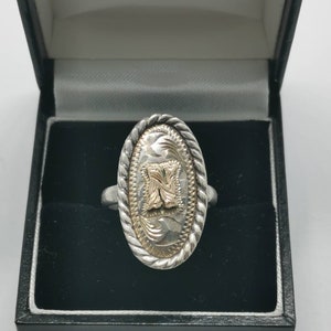 Puede incluir: Un anillo ovalado de plata y oro con un diseño de cuerda alrededor del borde. El anillo tiene un diseño de oro en el centro.