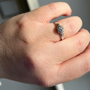 Puede incluir: Un anillo de plata con un grupo de peque&ntilde;as piedras redondas y transparentes dispuestas en un patr&oacute;n circular. El anillo est&aacute; en un dedo, y la mano est&aacute; ligeramente desenfocada contra un fondo brillante.