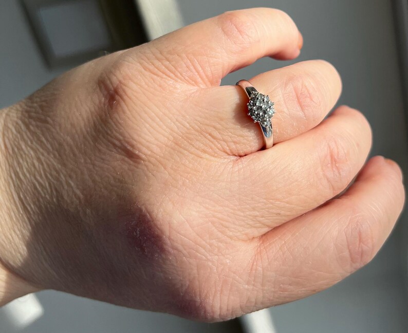 Puede incluir: Un anillo de plata con un grupo de peque&ntilde;as piedras redondas y transparentes dispuestas en un patr&oacute;n circular. El anillo est&aacute; en un dedo, y la mano est&aacute; ligeramente desenfocada contra un fondo brillante.