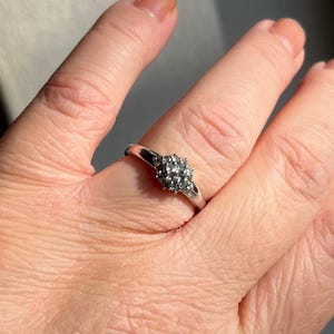 Puede incluir: Un anillo de plata con un grupo de peque&ntilde;as piedras claras dispuestas en un patr&oacute;n circular. El anillo est&aacute; en un dedo, y la banda es delgada y pulida. Las piedras brillan a la luz, creando una apariencia delicada y elegante.