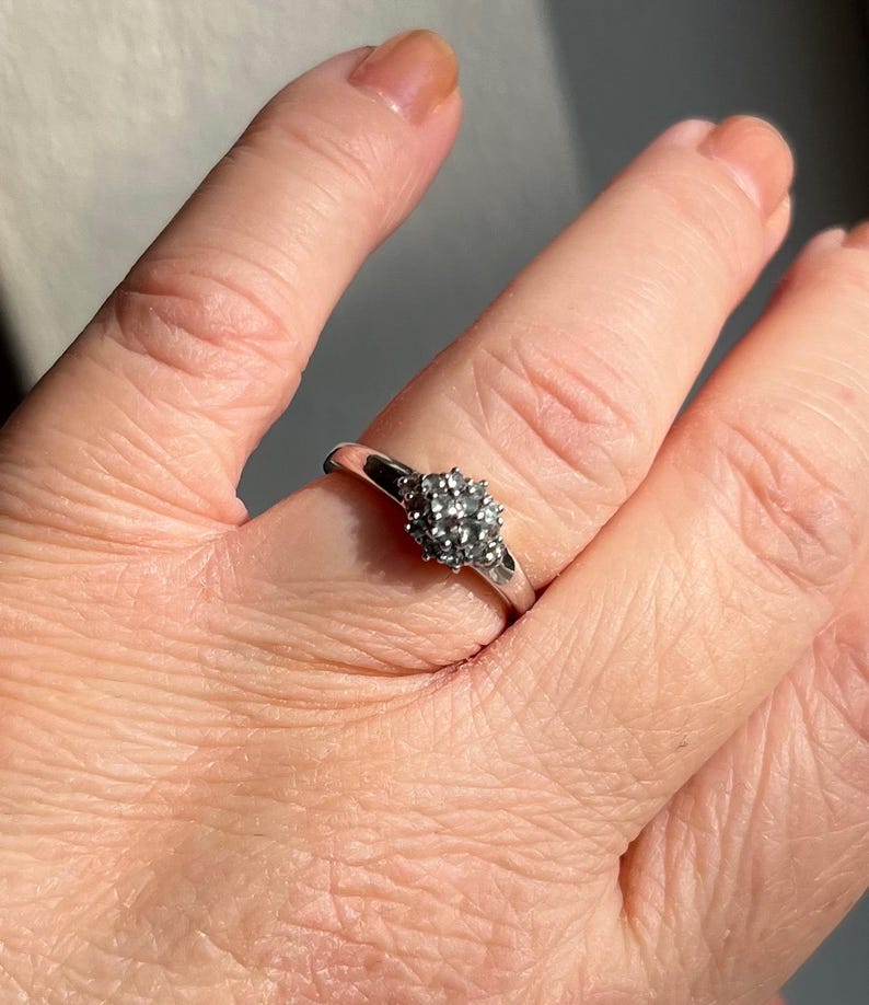 Puede incluir: Un anillo de plata con un grupo de peque&ntilde;as piedras claras dispuestas en un patr&oacute;n circular. El anillo est&aacute; en un dedo, y la banda es delgada y pulida. Las piedras brillan a la luz, creando una apariencia delicada y elegante.