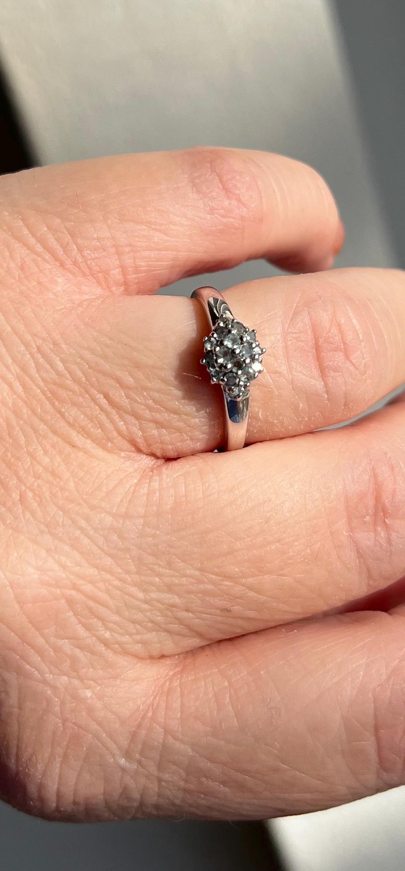 Puede incluir: Un anillo de plata con un grupo de peque&ntilde;as piedras preciosas redondas y transparentes. La banda del anillo es lisa y pulida, y las piedras est&aacute;n engastadas juntas, creando un efecto brillante. El anillo se muestra en un dedo.