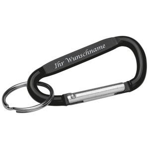 May include: Black carabiner clip with a silver key ring. The clip has the text "Ihr Wunschname" in white script. The carabiner is made of metal and has a spring-loaded gate for secure closure.