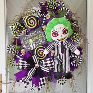 May include: A Halloween wreath featuring a green and black striped character with a green face and a black tie. The wreath is decorated with purple, black, and white ribbon, green and black candy cane shapes, and a sign that reads "It's Showtime".