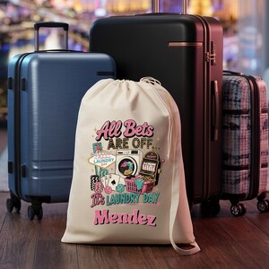 May include: A cream-colored drawstring laundry bag featuring a graphic print with a washing machine, slot machine, playing cards, and the text All Bets Are Off It's Laundry Day Mendez, positioned in front of three suitcases of varying sizes.