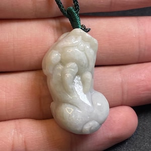 Op de afbeelding: Een witte jade hanger gegraveerd in de vorm van een mythisch wezen, hangend aan een groen koord. De hanger is gedetailleerd met ingewikkelde gravures en heeft een gladde, gepolijste afwerking.