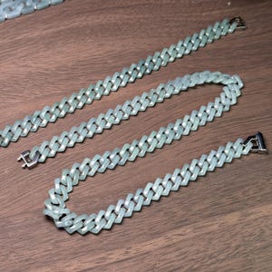 May include: A light green jade necklace and bracelet set. The jewelry features a curb chain design with a silver clasp. The pieces are displayed on a wooden surface, showcasing the cool tones of the jade.