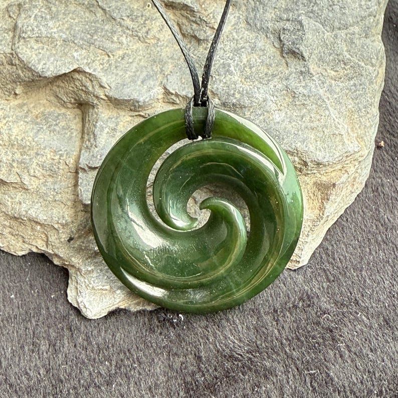 May include: A green jade pendant with a spiral design, hanging on a black cord. The pendant is a traditional Maori symbol called a Koru, representing new beginnings and growth.
