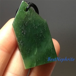May include: A dark green jade pendant with a smooth, polished surface. The pendant is shaped like a house and has a hole at the top for a cord. The pendant is hanging from a black cord.