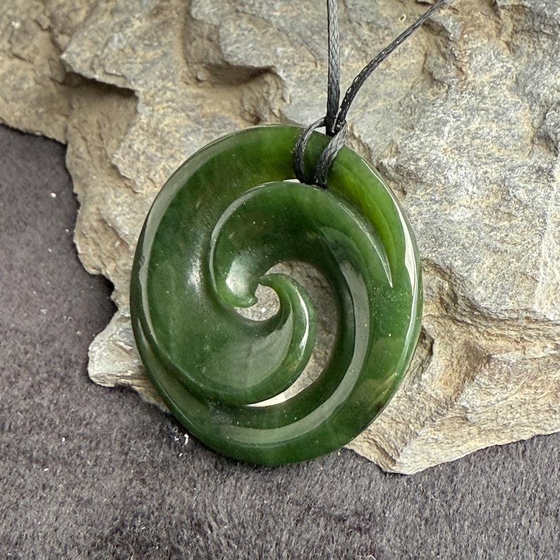 May include: A green jade pendant with a spiral design, known as a Maori Koru, on a black cord.