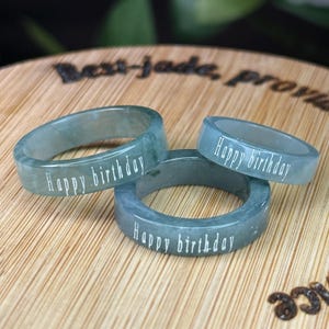Puede incluir: Tres anillos de jade en tonos verdes y azules. Cada anillo tiene la inscripción "Happy birthday" en blanco. Los anillos están sobre una superficie de madera.