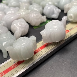 Könnte beinhalten: Eine Sammlung kleiner, geschnitzter Jadeit-Teekannen in verschiedenen Weiß- und Grüntönen. Die Teekannen sind aufwendig detailliert, mit Ausgüssen, Griffen und Deckeln. Ein Lineal mit Millimeter-Markierungen befindet sich im Vordergrund.