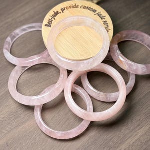 May include: A collection of rose quartz bangles in shades of pink are displayed. The circular bracelets have a smooth, polished finish. A wooden disc with text is in the background. The bangles are made of a translucent material.