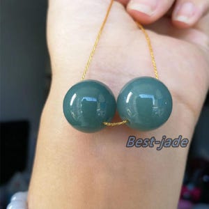 Puede incluir: Dos esferas de jade de color verde azulado ensartadas en un cordón dorado. Las esferas son lisas y reflectantes, con el texto "Best-jade" visible a la derecha. Las esferas se sostienen en una mano.