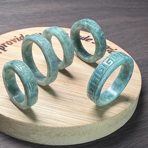 Puede incluir: Cinco anillos de jade con un diseño geométrico tallado se exhiben sobre una superficie de madera. Los anillos son de color verde pálido y tienen un acabado liso y pulido. Los anillos son de diferentes tamaños y están dispuestos de forma atractiva.