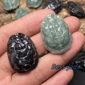 May include: Two jade turtle carvings, one green and one black, are held in a hand. The green turtle has a detailed shell pattern. The black turtle has a more mottled appearance.