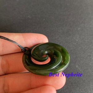 May include: A green jade pendant with a spiral design, known as a Maori Koru, on a black cord. The pendant is carved from New Zealand nephrite.