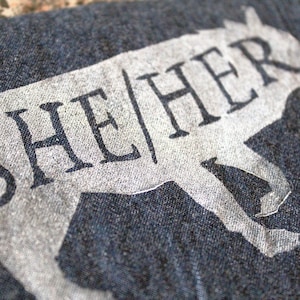 Op de afbeelding: Een donkergrijs T-shirt met een wit silhouet van een lopend dier en de tekst "SHE/HER" erop gedrukt.