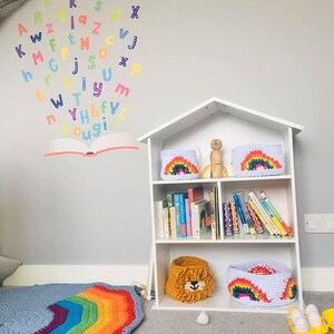 May include: A white bookshelf with books and decorative crochet baskets. The baskets feature rainbow and lion designs. A rainbow-colored crochet rug and wall decals of letters and a book complete the playful nursery decor.