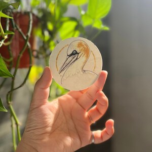 Peut inclure: Un autocollant rond avec un fond blanc et une illustration dessinée à la main d'un oiseau de la famille des fous de bassan avec un bec jaune et des yeux noirs. L'oiseau est entouré de deux cercles concentriques de couleur dorée.