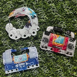 Peut inclure: Trois porte-clés en acrylique sur le thème des Transformers. L'un est en forme de nuage blanc avec des jets colorés, un autre est un boombox rouge et blanc, et le dernier est une cassette bleue. Le tout sur un fond d'herbe verte.