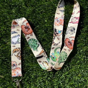 May include: A white lanyard with a repeating pattern of cartoon geckos and snakes. The lanyard has a silver clip on one end.
