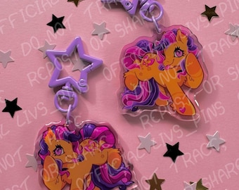 Op de afbeelding: Twee sleutelhangers met tekenfilmpony's met oranje en paarse manen en staarten. Elke sleutelhanger heeft een stervormige sluiting. De achtergrond is roze met stervormige confetti en de tekst "OFFICIAL LIVS PINS OF".