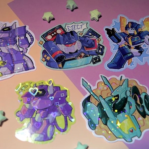 May include: A selection of colourful, die-cut stickers featuring cartoon robots. The stickers depict various robot designs in shades of purple, blue, and teal, with some holographic accents. The stickers are arranged on a pink and purple background, with star-shaped decorations.