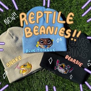 Puede incluir: Tres gorros con bordados con temática de reptiles se muestran sobre hierba verde. Los gorros son de color azul claro, beige y negro. El gorro azul claro dice "Reptile Beanies!!" y tiene un lagarto de lengua azul. El gorro beige dice "Snake" y tiene una serpiente. El gorro negro dice "Beardie" y tiene un lagarto.